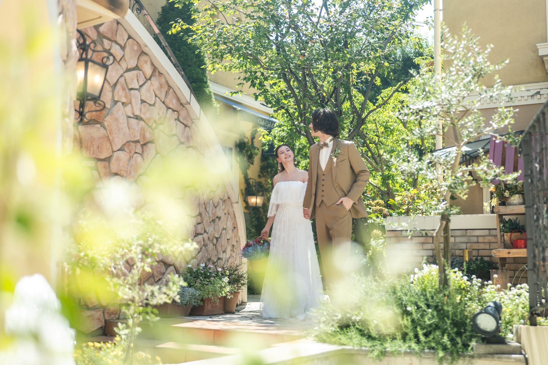 アネーリ大宮の自然光溢れるガーデンを歩く新郎新婦。ヨーロッパ風の石積みの壁と豊かな緑に囲まれた、さいたま市で叶うナチュラルなガーデンウェディングのロケーションフォト。
