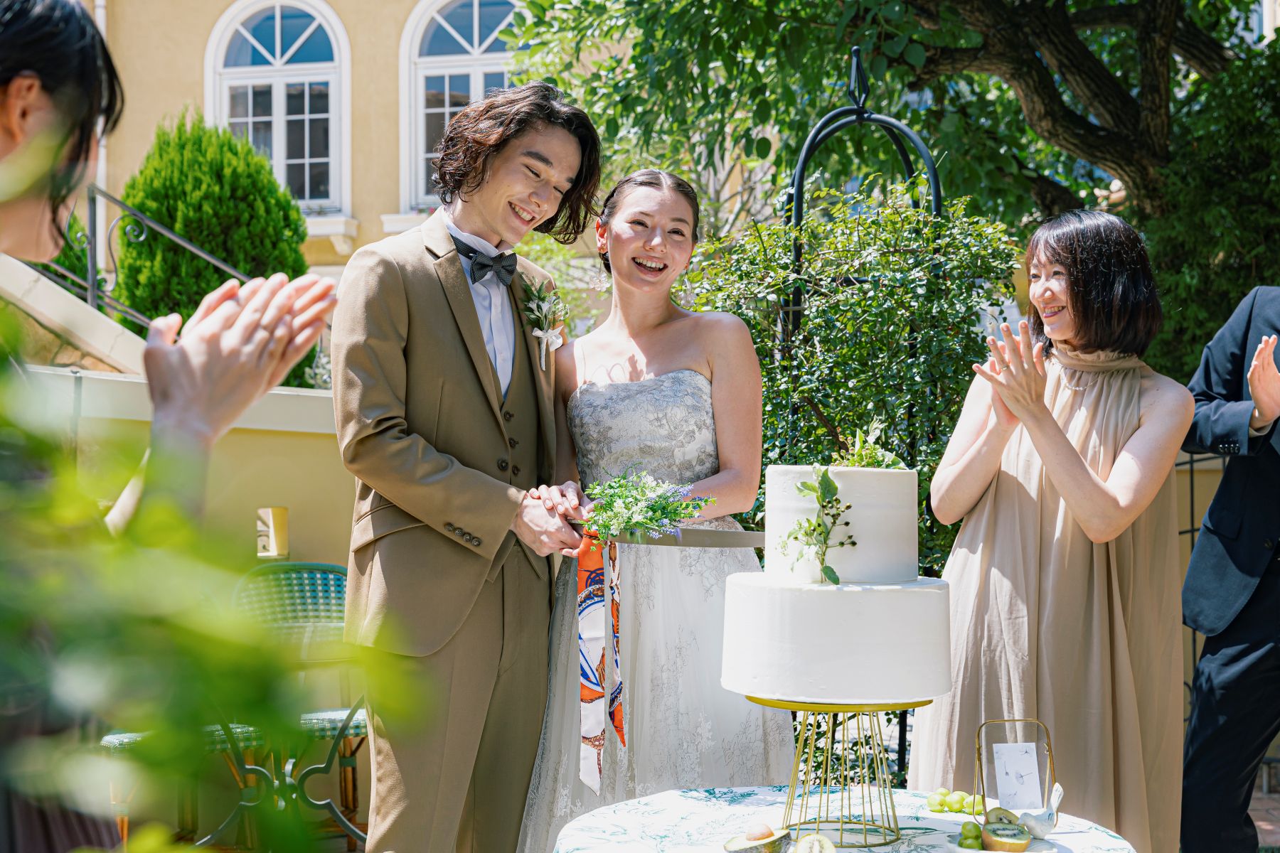 さいたま市の結婚式場「アネーリ大宮」のガーデンウェディング。青空の下、南欧風の邸宅を背景にした屋外テラスで、ゲストの温かい拍手に包まれながら笑顔でウェディングケーキ入刀を行う新郎新婦。開放感と幸福感あふれる屋外演出のシャッターチャンス。