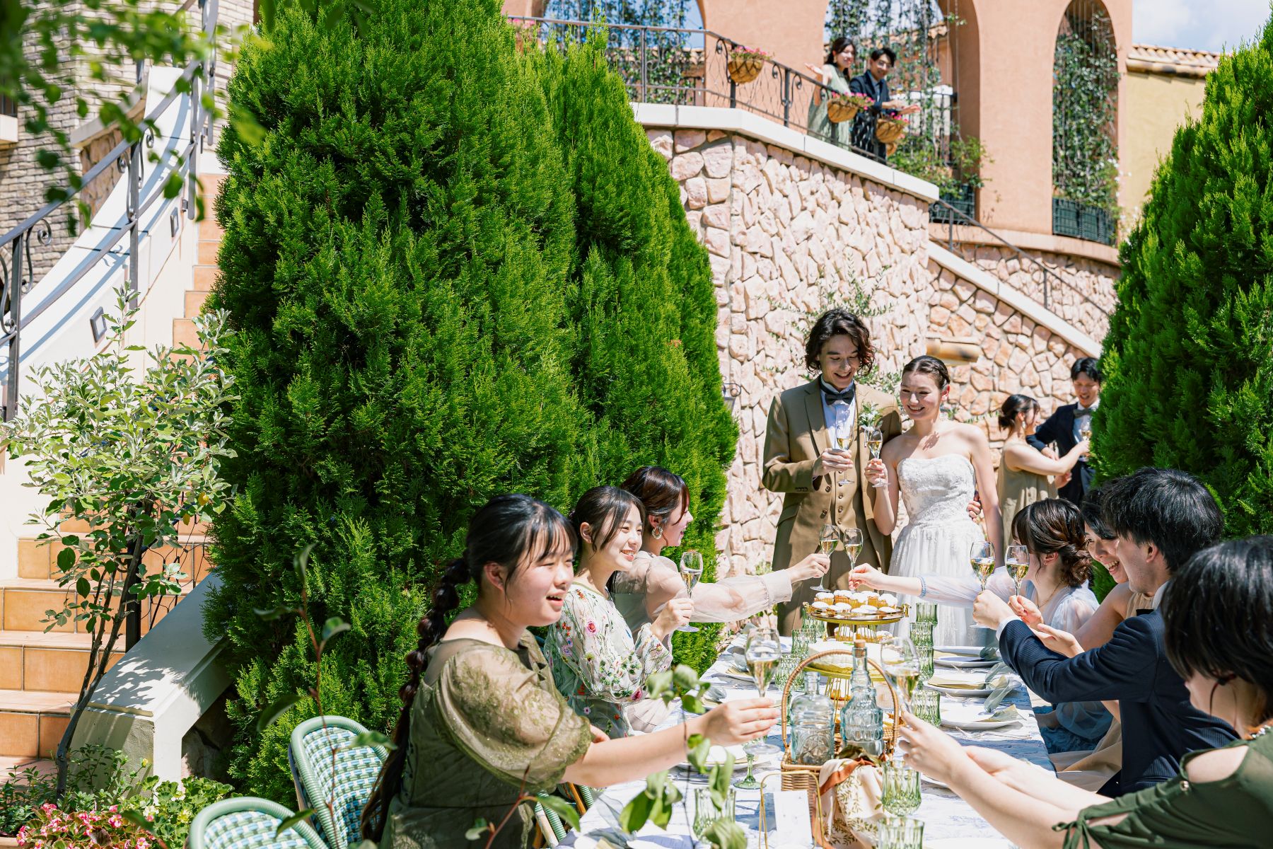 大宮の結婚式場「アネーリ大宮」のガーデンウェディング。青空と豊かな緑に囲まれた屋外テラスで、新郎新婦とゲストが笑顔で乾杯を交わす開放感あふれるパーティーの様子。ヨーロッパのリゾートを彷彿とさせる石積みの邸宅を背景にした、アットホームな屋外演出。