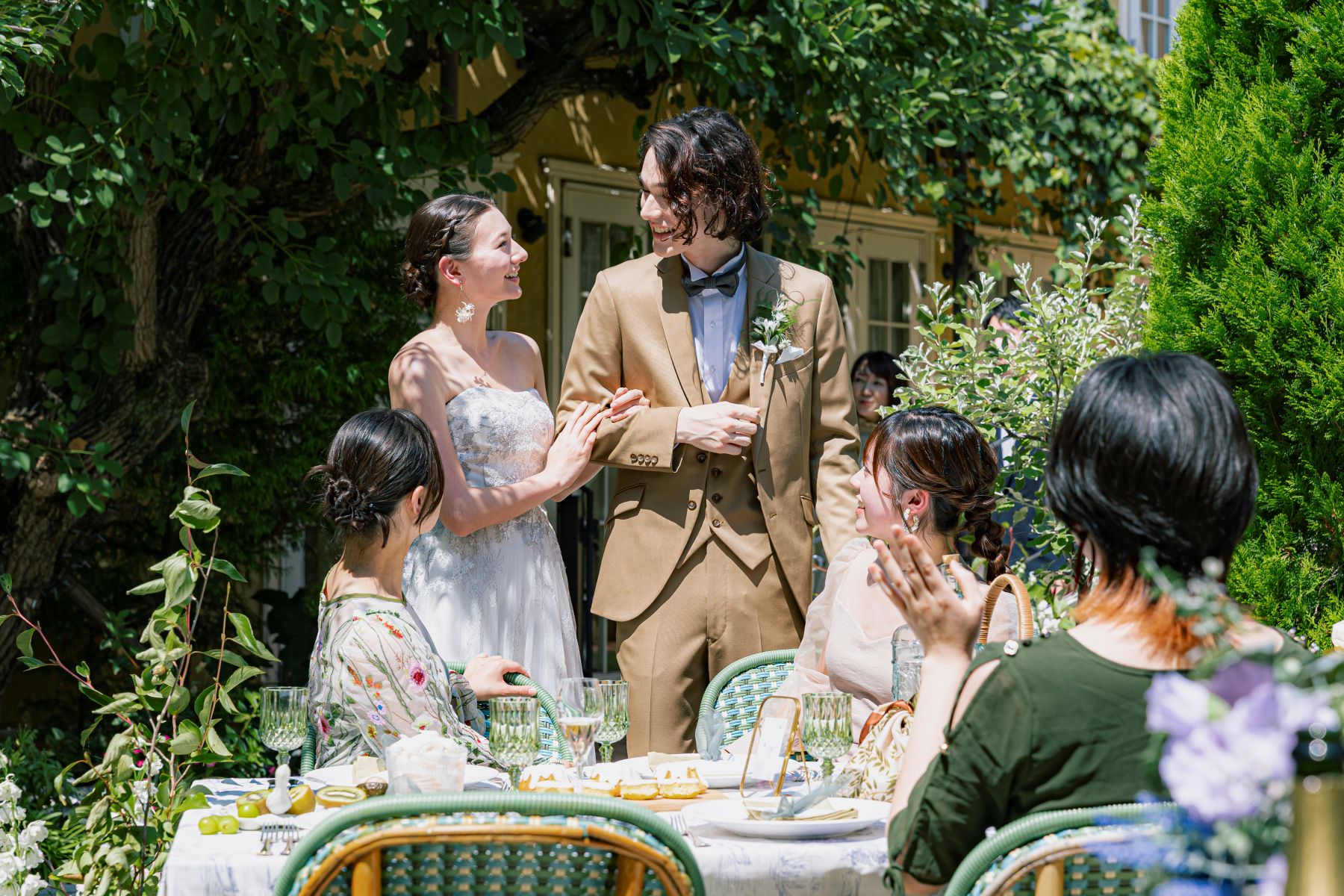 さいたま市の結婚式場「アネーリ大宮」のガーデンウェディング。豊かな緑と大きな樹木に囲まれた屋外テラスで、新郎新婦がゲストと親密に語らうアットホームな歓談シーン。自然光が降り注ぐ中での開放的なパーティー演出の様子。