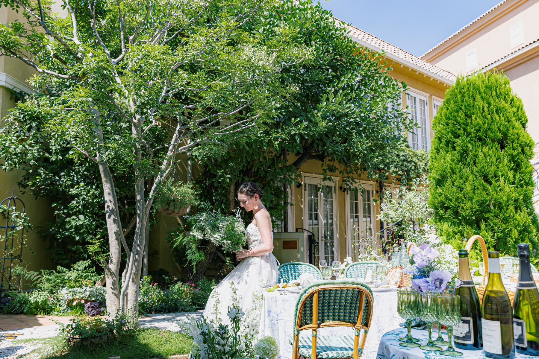 さいたま市の結婚式場「アネーリ大宮」のプライベートガーデン。大きなシンボルツリーと邸宅を背景にした開放感あふれる屋外テラスで、自然光を浴びながらブーケを手に佇む花嫁。南欧のリゾート邸宅のような雰囲気の中で叶う、ナチュラルで上質なガーデンウェディングの様子。