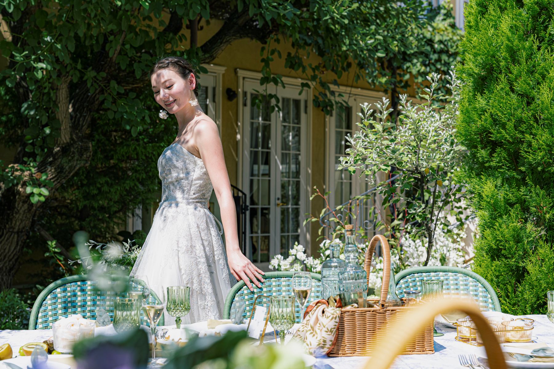 さいたま市の結婚式場「アネーリ大宮」のガーデンウェディング。自然光が降り注ぐ屋外テラスで、繊細な刺繍が施されたウェディングドレスを纏った花嫁が、緑のグラスやバスケットで彩られたゲストテーブルの傍らに佇む様子。緑豊かな庭園と邸宅を背景にした、ナチュラルで開放的なパーティー風景。
