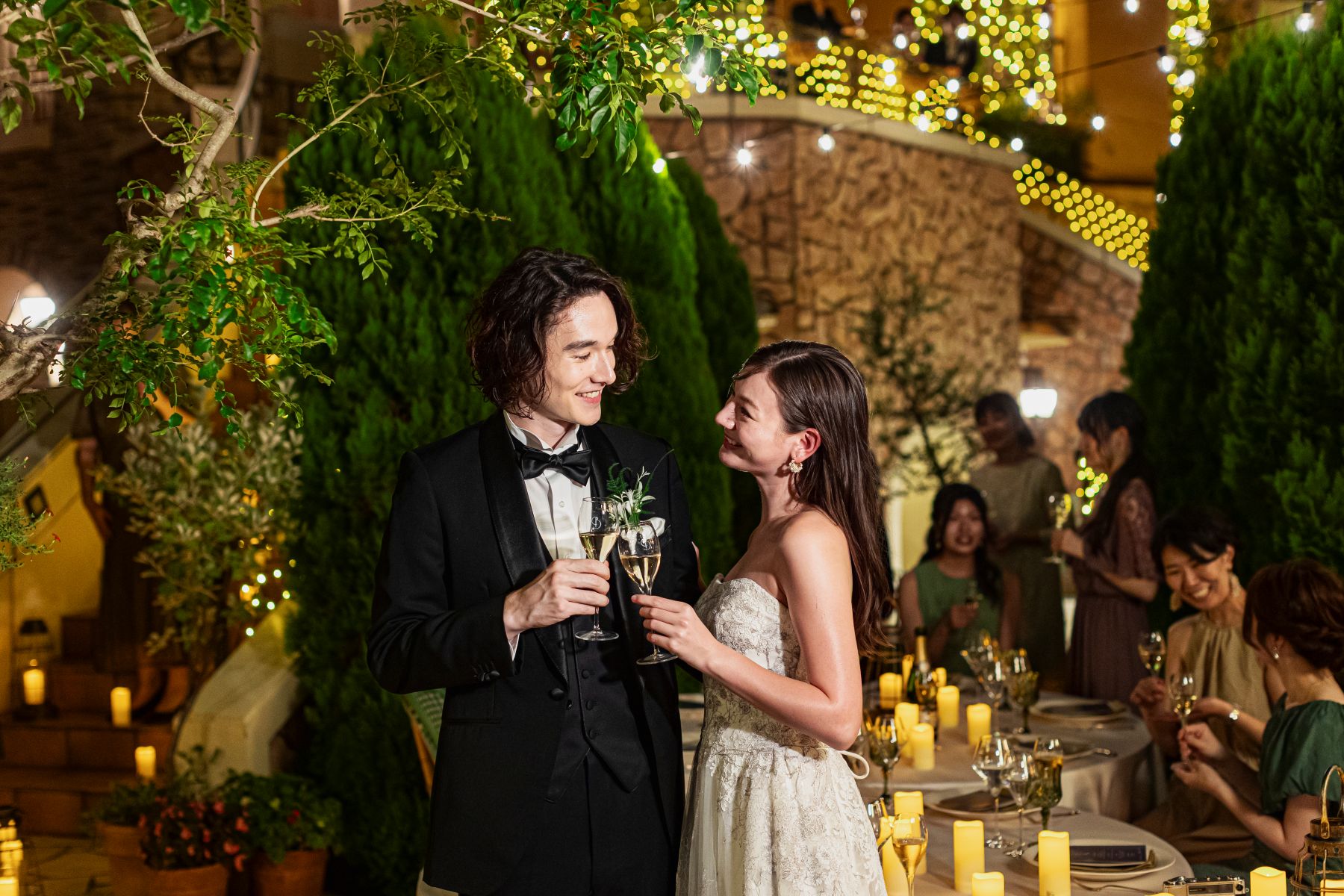 さいたま市の結婚式場「アネーリ大宮」での幻想的なナイトウェディング。暖かなキャンドルの光とイルミネーションが輝く夜のガーデンテラスで、シャンパングラスを手に微笑み合う新郎新婦。ゲストに囲まれたロマンチックで上質な屋外パーティーの様子。