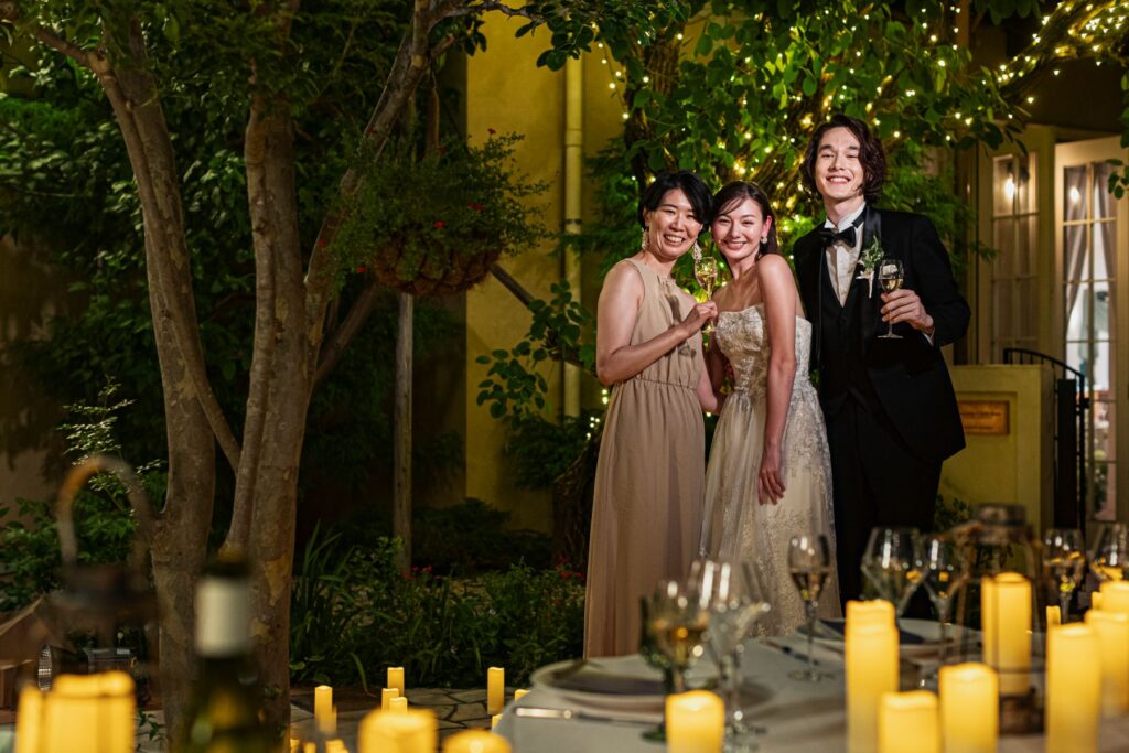 【さいたま市結婚式場アネーリ大宮】のライトアップされたガーデンにて、ゲストと笑顔で乾杯を交わす新郎新婦様。さいたま市にある市の中心部で叶える、開放感あふれる夜のパーティ空間を詳細に描写しており、さいたま市内での結婚式らしい洗練された非日常的なおもてなしを表現しています。