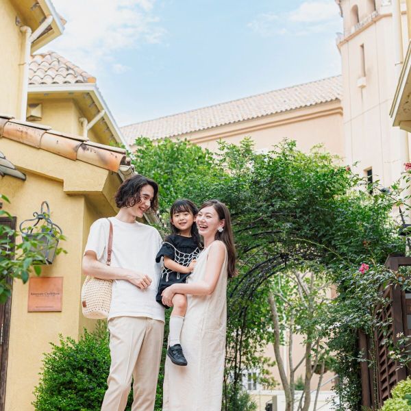 さいたま市の結婚式場「アネーリ大宮」で行われる結婚3周年記念式「ノス・ド・フロマン」。青空と緑、南欧風の街並みを思わせる邸宅をバックに、お子様を抱っこして笑顔で語らう家族3人の幸せな様子。結婚式後も家族の絆を育むアニバーサリーイベントのワンシーン。