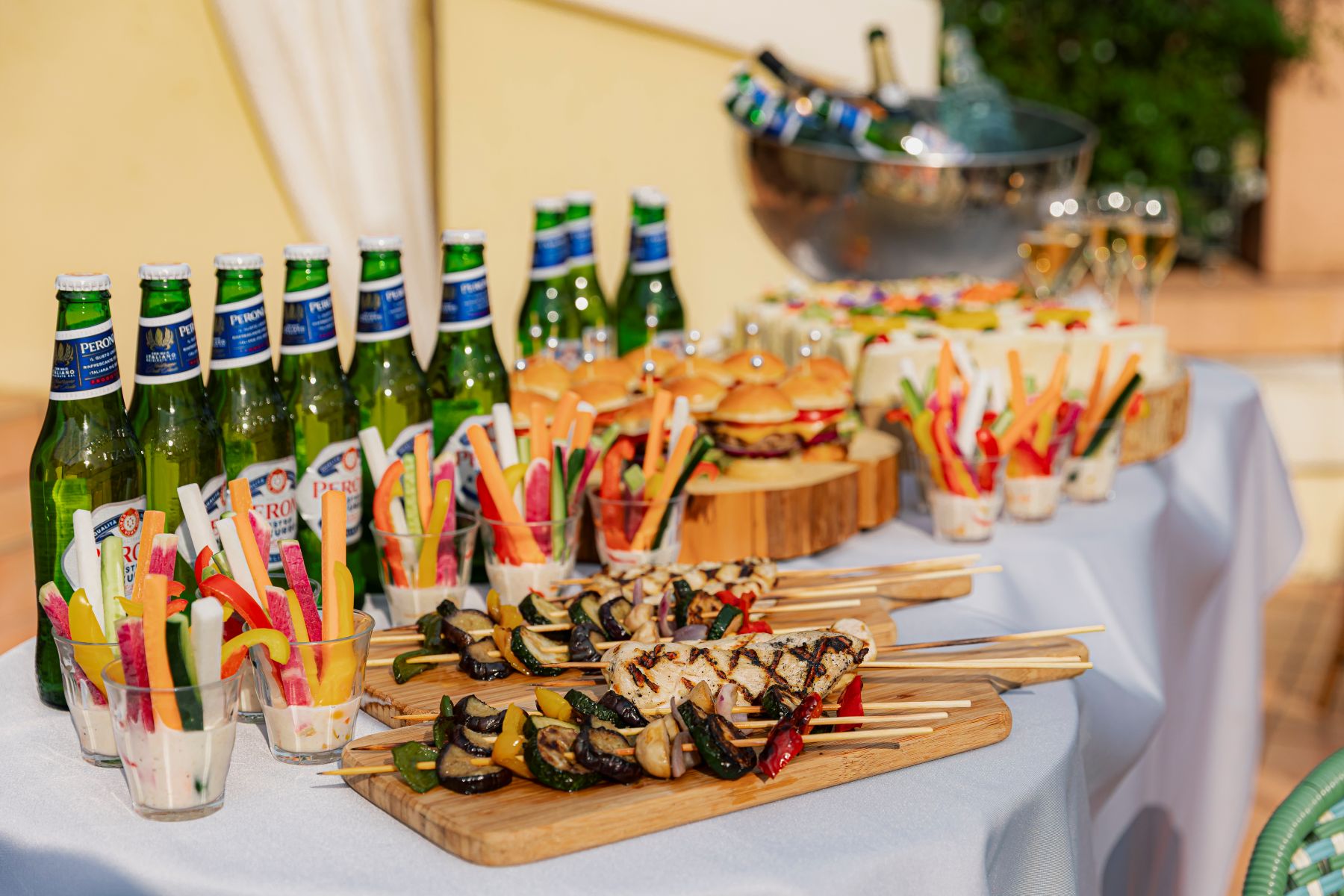 さいたま市の結婚式場「アネーリ大宮」のガーデンウェディングで振る舞われる婚礼料理。開放的な屋外テラスでのパーティーを彩る、香ばしく焼き上げた串焼きグリルやミニバーガー、フレッシュな野菜スティックなどのビュッフェスタイルのメニューと、冷えたビールのコーディネート。
