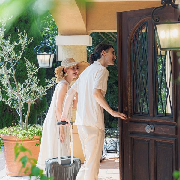 さいたま市の結婚式場「アネーリ大宮」で叶える結婚3周年記念式「ノス・ド・フロマン」。リゾートを訪れたような旅の装いで、キャリーケースを手に重厚な邸宅の扉を開ける夫婦。自然光が差し込むエントランスで、特別なアニバーサリーの始まりを予感させる一枚。