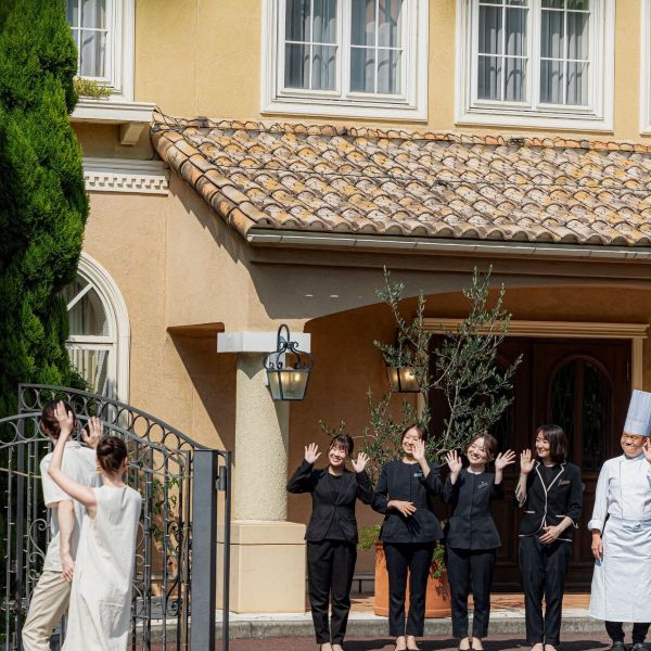 さいたま市の結婚式場「アネーリ大宮」の結婚3周年記念式『ノス・ド・フロマン』を終えた夫婦を、笑顔で見送るスタッフ一同。南欧風の邸宅を背景に、プランナーやシェフが手を振る温かなおもてなしの風景。挙式後も「いつでも帰ってこられる場所」として家族に寄り添い続ける会場の魅力を伝える一枚。