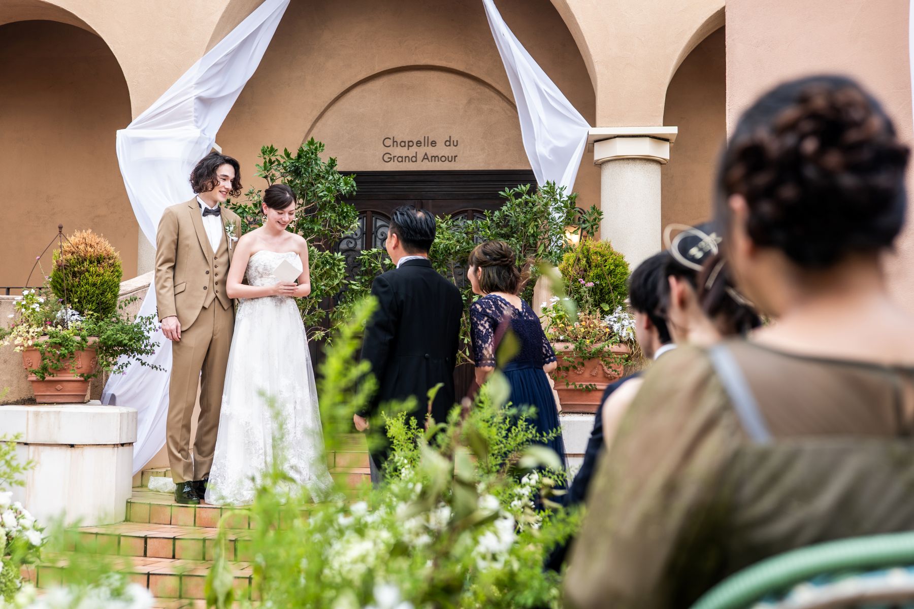 「大宮の結婚式場『アネーリ大宮』の開放的なガーデン人前式。緑豊かな屋外チャペルを背景に、ゲストの温かな祝福に包まれながら、新婦が手紙や誓いの言葉を読み上げる感動的なシーン。ナチュラルでアットホームな屋外ウェディングの魅力を伝える一枚。」