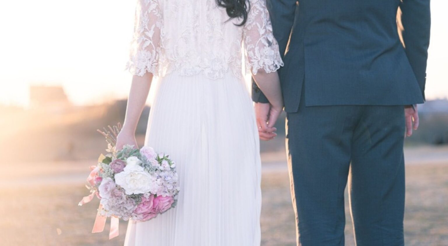 【さいたま市結婚式場アネーリ大宮】の広大なロケーションにて、夕暮れ時の光を浴びながら手を繋ぐウェディングドレス姿の新婦様と新郎様。さいたま市にある市の式場で叶える、ドラマチックな前撮りを詳細に描写しており、さいたま市内での結婚式らしい、情緒溢れる美しい一瞬を表現しています。