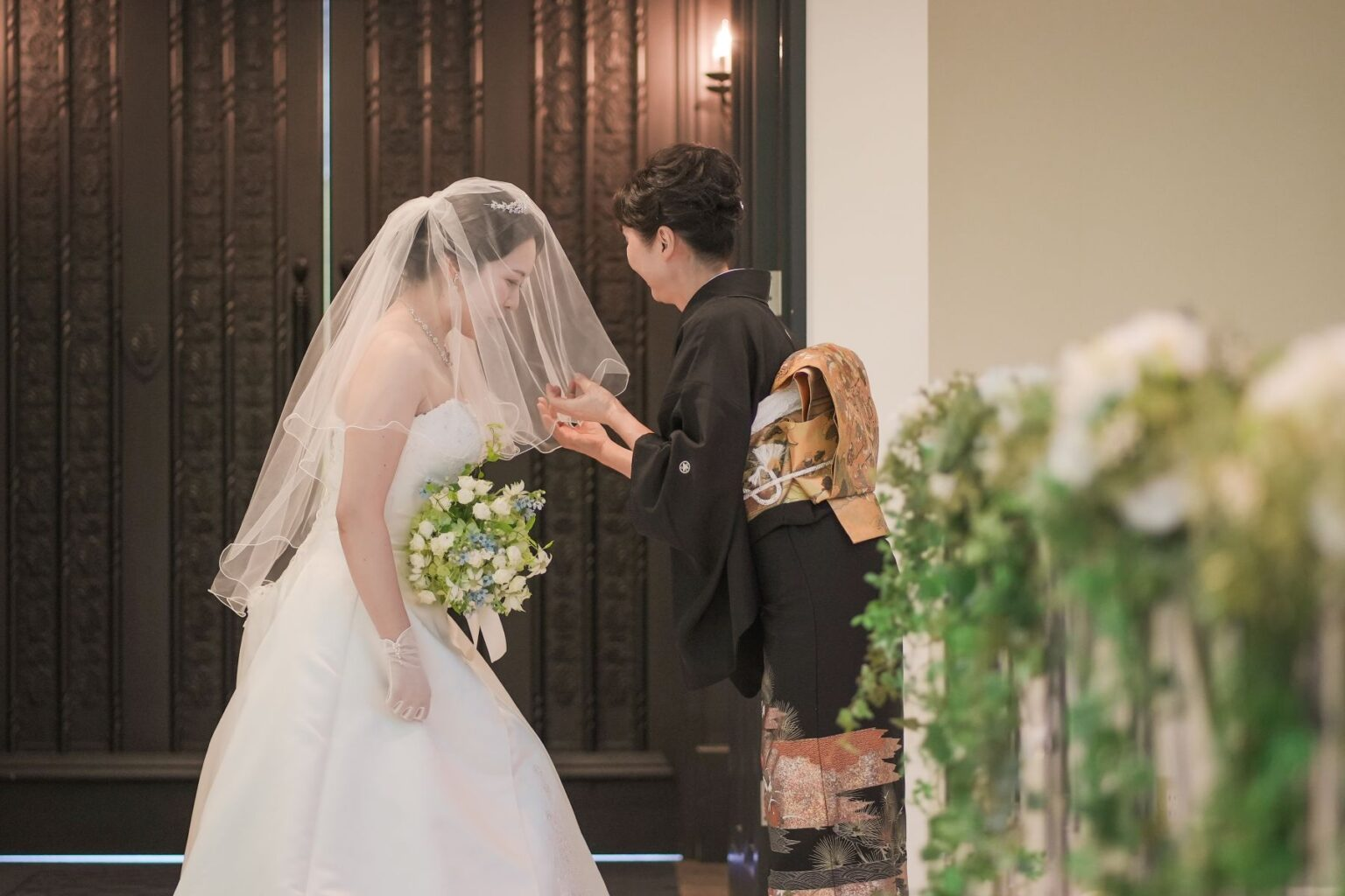 【さいたま市結婚式場アネーリ大宮】の開放的な屋外にて、長いベールに包まれながら静かに誓いのキスを交わす新郎新婦様。さいたま市にある市の式場で叶える、ベールダウンから続く神聖なセレモニーを詳細に描写しており、さいたま市内での結婚式らしい、深い愛と感動に満ちたウェディングシーンを表現しています。