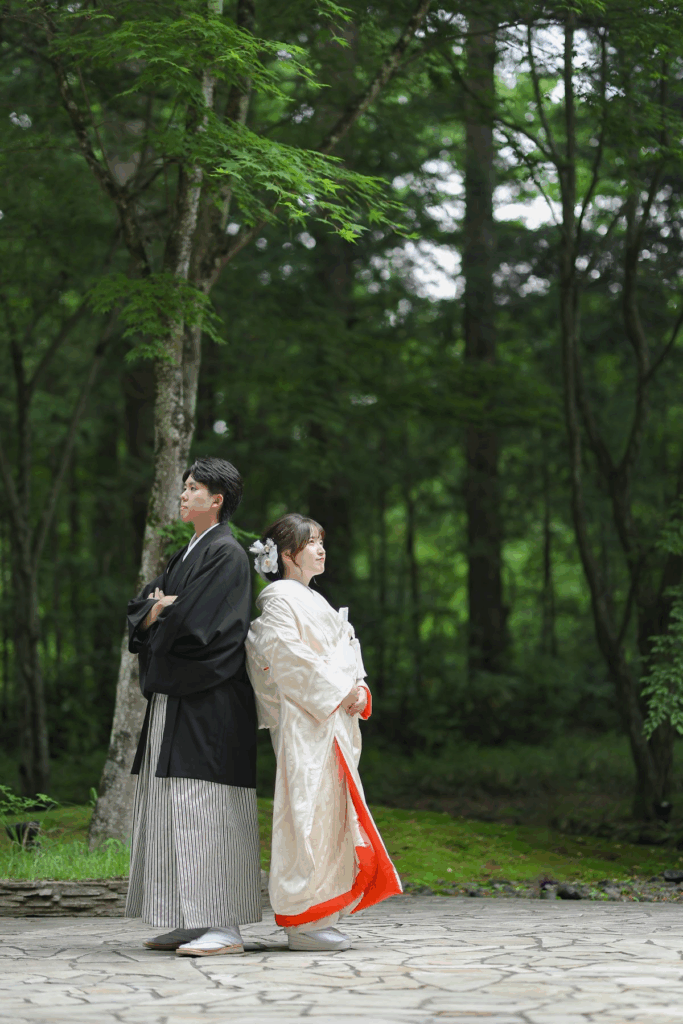 【さいたま市結婚式場アネーリ大宮】の豊かな自然に囲まれたロケーションで、背中合わせに立つ紋付袴姿の新郎様と白無垢姿の新婦様。さいたま市にある市の式場で叶える、新緑が美しい屋外フォトを詳細に描写しており、さいたま市内での結婚式を象徴するような、清らかで静謐な空気感を表現しています。