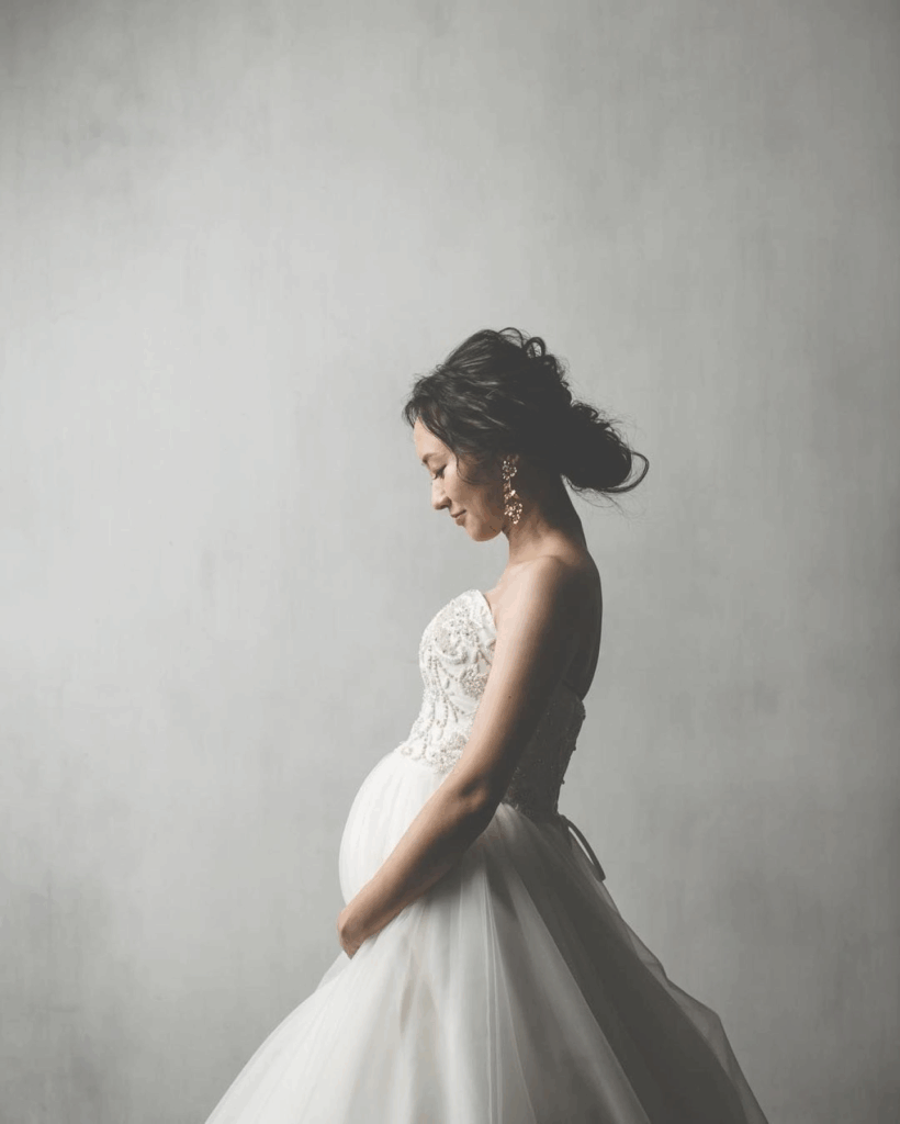 【さいたま市結婚式場アネーリ大宮】の柔らかな光の中で、お腹を優しく愛おしむマタニティ姿の花嫁様。さいたま市にある市の式場で叶える、神秘的で美しい瞬間を詳細に描写しており、さいたま市内での結婚式がお二人と赤ちゃんにとって最高の門出となるよう願いを込めています。