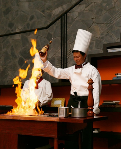 【さいたま市結婚式場アネーリ大宮】の披露宴会場にて、大きな炎を上げるフランベ演出を披露するシェフ。さいたま市にある市のバンケットで叶える、五感に響く美食のパフォーマンスを詳細に描写しており、さいたま市内での結婚式らしいダイナミックで記憶に残るおもてなしの瞬間を表現しています。