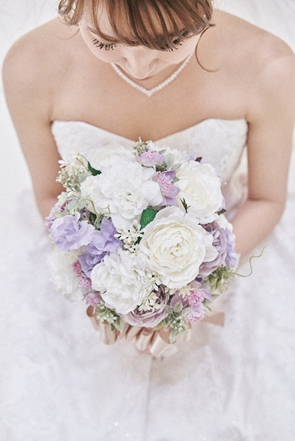 【さいたま市結婚式場アネーリ大宮】の光に包まれた空間で、白や紫の花々を束ねたブーケを大切に抱える花嫁様。さいたま市にある市の式場で叶える、純白のドレスと繊細なレースが調和した美しいソロショットは、さいたま市内での結婚式を象徴するような無垢で幸せな瞬間を詳細に描写しています。