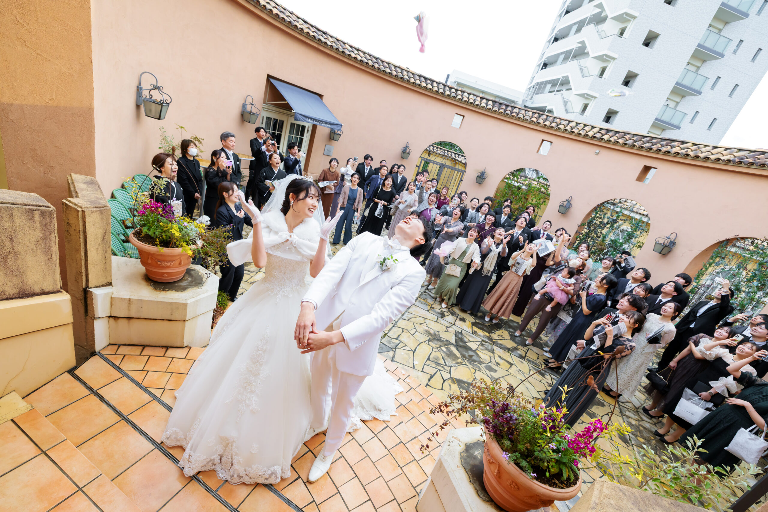 【さいたま市結婚式場アネーリ大宮】の南欧風の広々とした中庭にて、ゲストの皆様に見守られながらブーケトスを行う新郎新婦様。さいたま市にある市の式場で叶える、笑顔と活気に満ちた屋外演出は、さいたま市内外から集まった大切な方々との喜びの瞬間を詳細に描写しています。