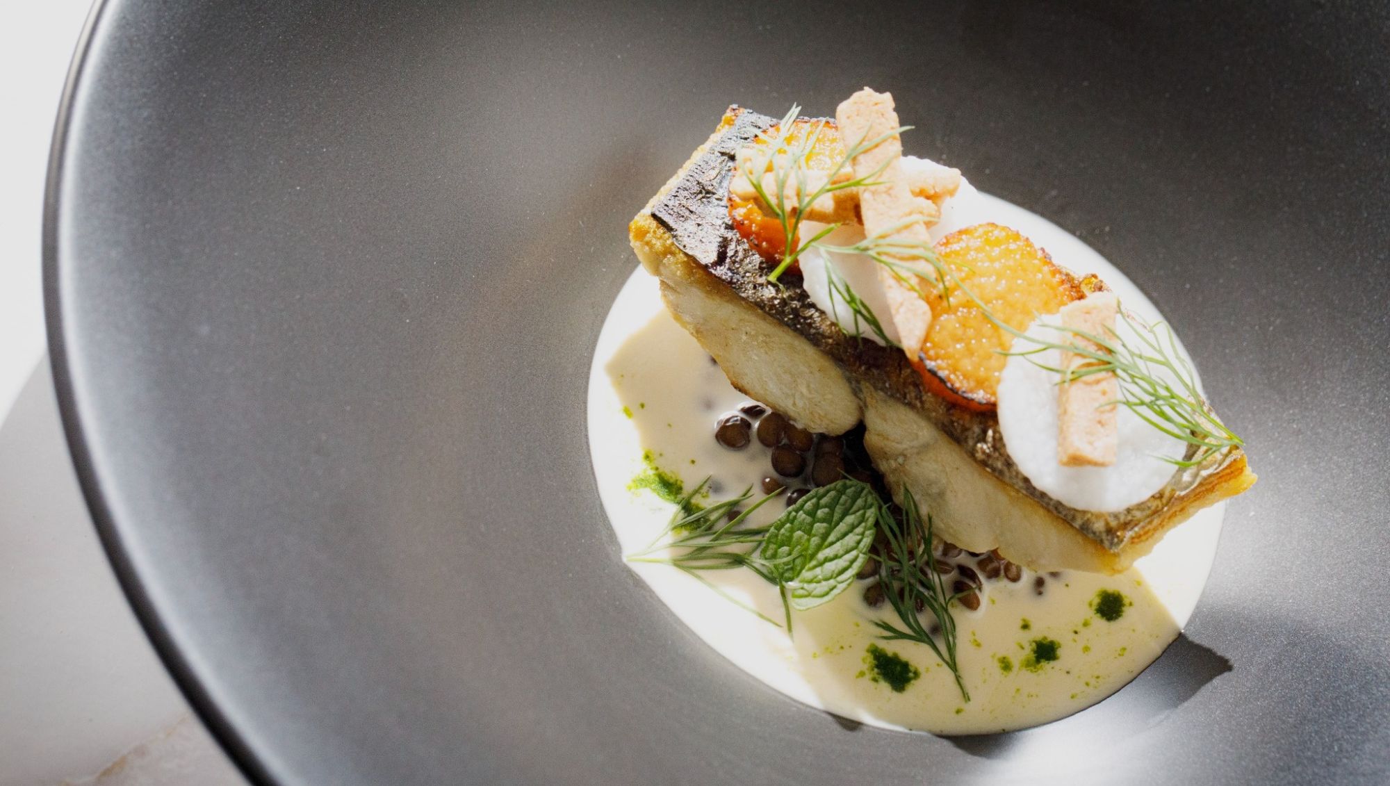 アネーリ大宮の婚礼料理。黒の深皿に盛り付けられた白身魚のポワレと、クリーミーなソース、爽やかなハーブのあしらい。大宮の結婚式場でゲストに振る舞われる、旬の素材を活かした本格フレンチの魚料理。